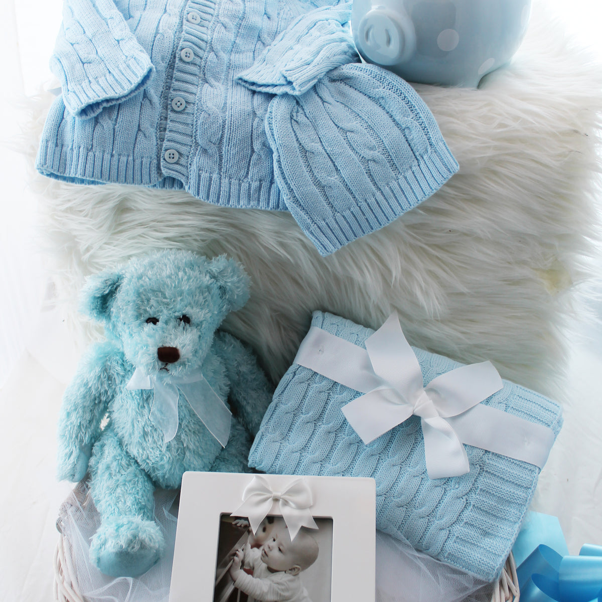 Hello Little One baby gift basket with sweater set, piggy bank, teddy bear, photo frame, and cable knit blanket in a wicker basket, shown in blue