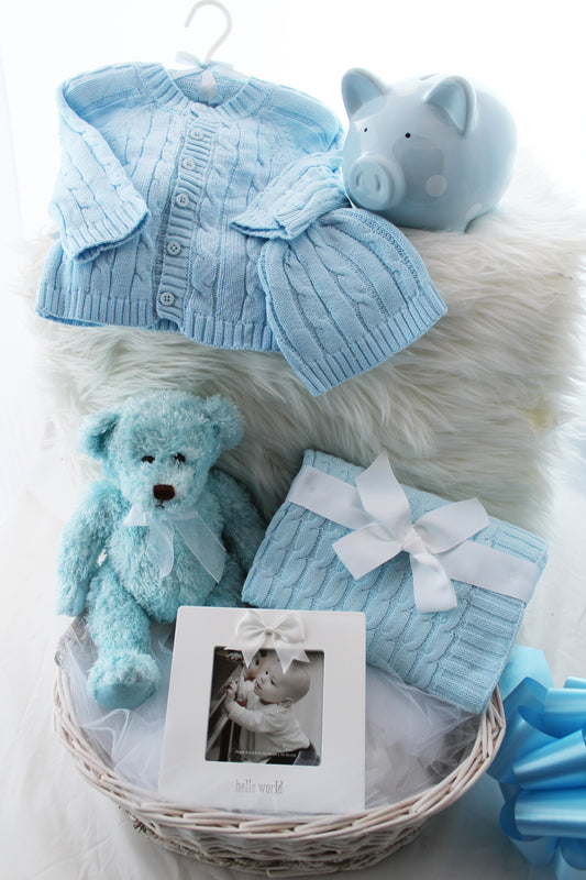 Hello Little One baby gift basket with sweater set, piggy bank, teddy bear, photo frame, and cable knit blanket in a wicker basket, shown in blue
