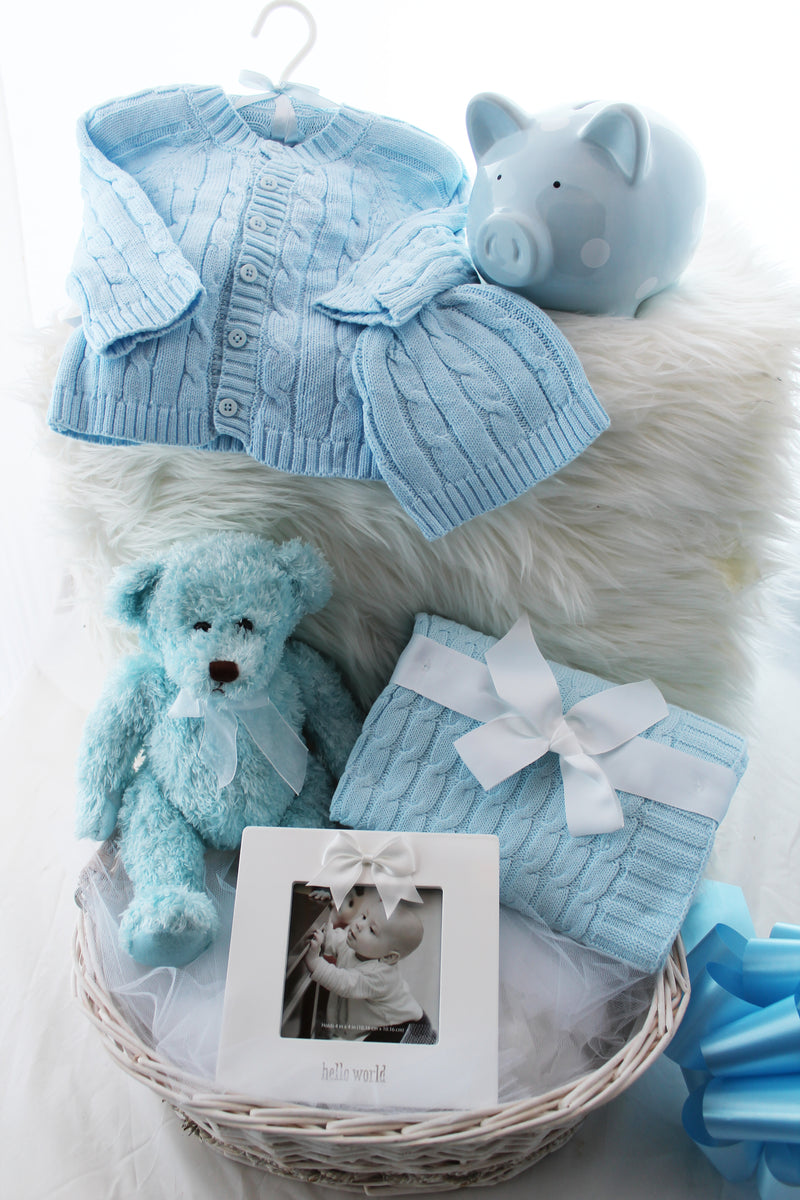 Hello Little One baby gift basket with sweater set, piggy bank, teddy bear, photo frame, and cable knit blanket in a wicker basket, shown in blue