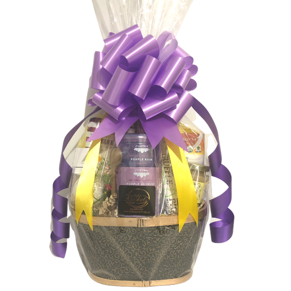 A calming gift basket featuring assorted teas, honey, a bath bomb, chocolate, and snacks, wrapped in cellophane with purple and yellow ribbons, and purple bow.